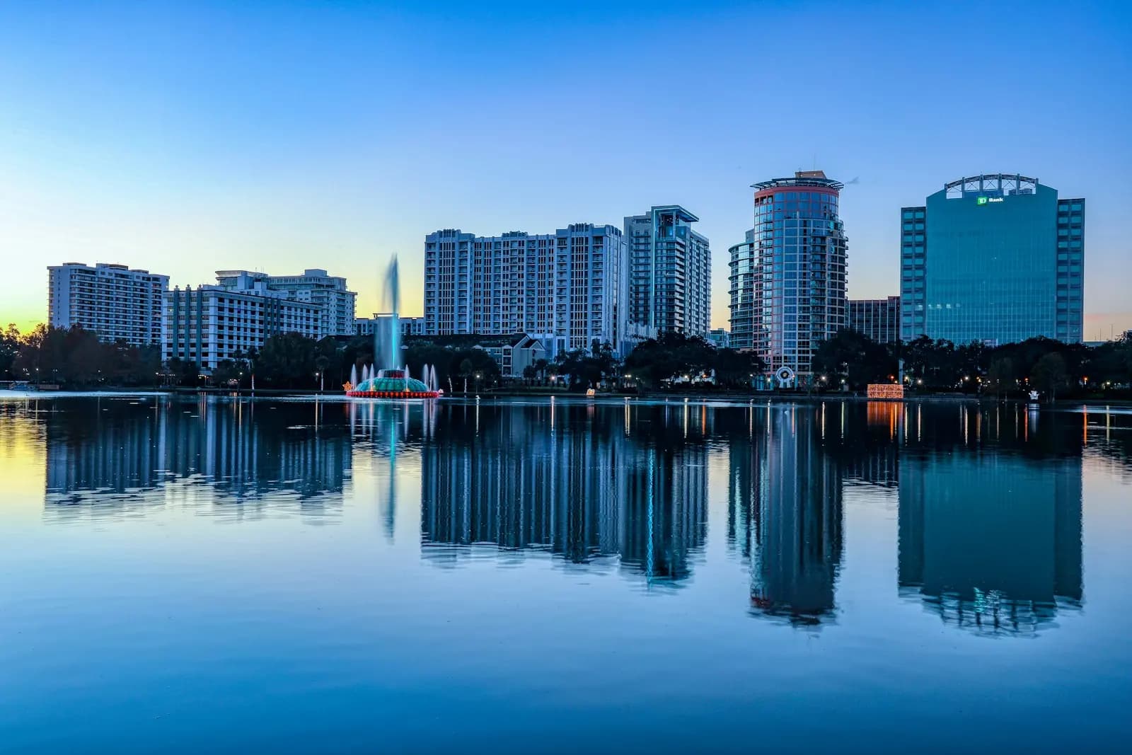 Alacakaranlıkta Orlando’da Lake Eola; gölün yüzeyinde yansıyan yüksek binalar ve ortadaki fıskiye.